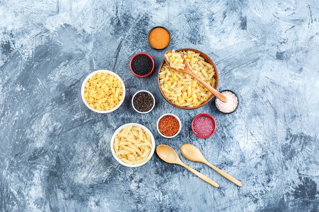 Raw pasta in bowls with spices, wooden spoons top view on a grey plaster background