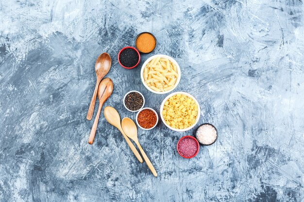 Pasta cruda in ciotole con spezie, vista dall'alto di cucchiai di legno su uno sfondo di gesso grigio