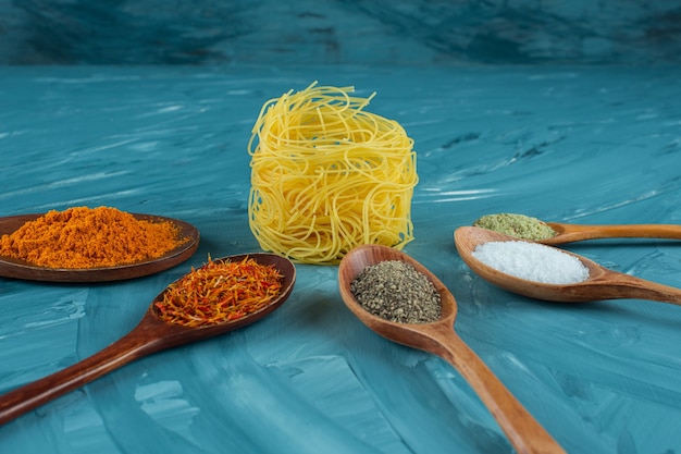 Raw pasta nest with spoons of condiments on blue surface