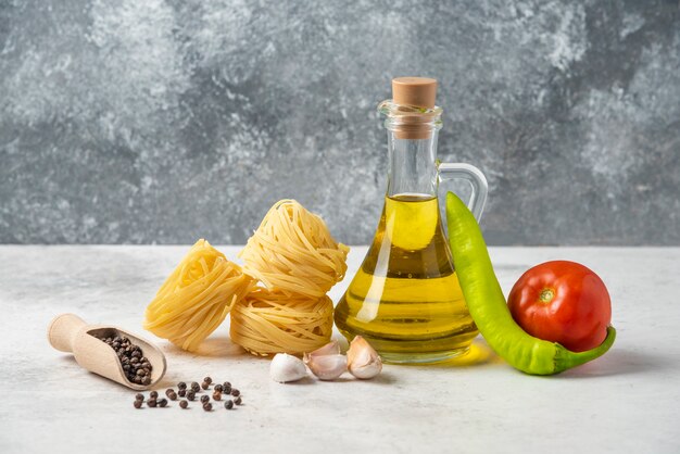 Raw pasta nests, bottle of olive oil, pepper grains and vegetables on white table. 