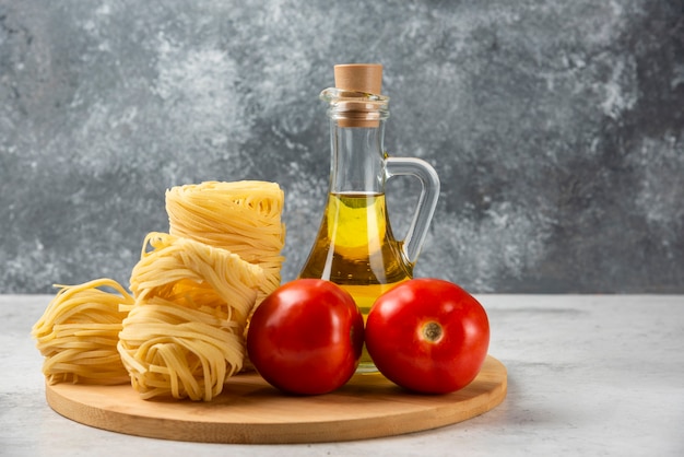 Raw pasta nests, bottle of olive oil and tomatoes on wooden plate. 