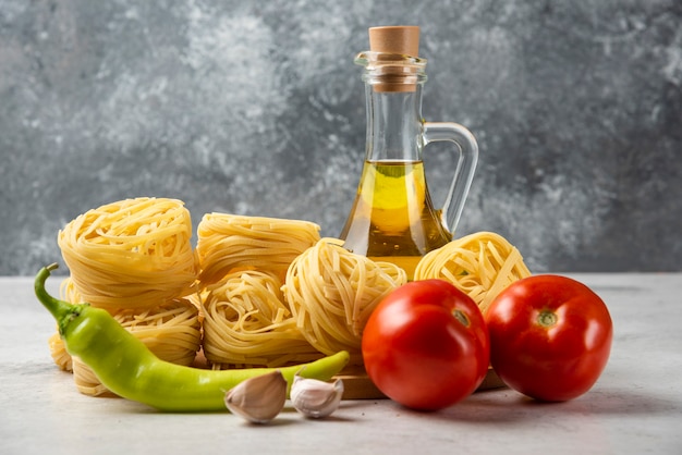 Raw pasta nests, bottle of olive oil and vegetables on white table. 