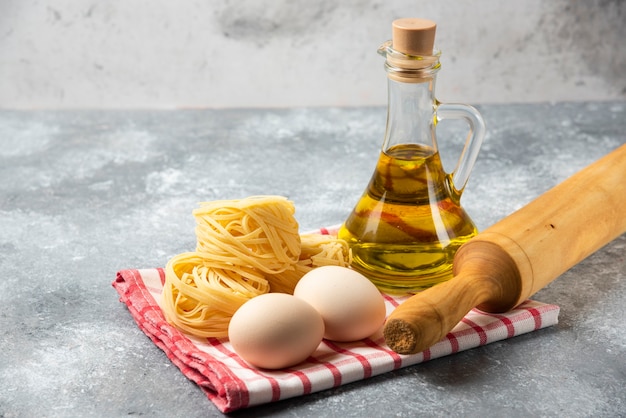 Raw pasta nests, eggs, bottle of olive oil and rolling pin on marble table. 