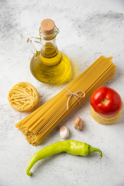 Raw pasta nests, spaghetti, bottle of olive oil and vegetables on white surface. 