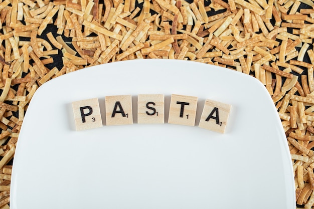 Raw pasta scattered around white plate with wooden letters. High quality photo