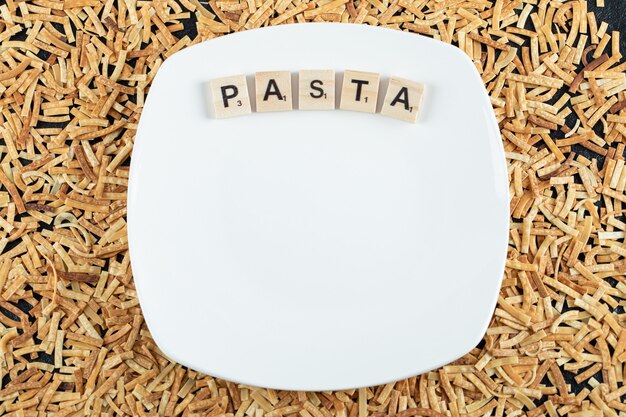 Raw pasta scattered around white plate with wooden letters.