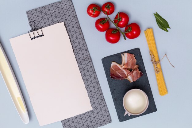 Raw pasta; tomatoes; meat; white sauce; bay leaves and blank white paper with napkin on plain backdrop