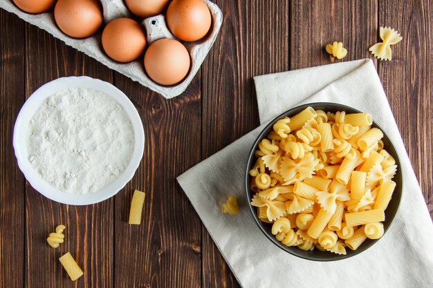 Raw pasta with eggs, flour in a bowl on wooden and kitchen towel, flat lay.