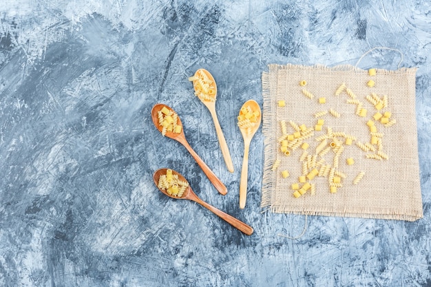 Raw pasta in wooden spoons on a plaster and piece of sack background. top view.