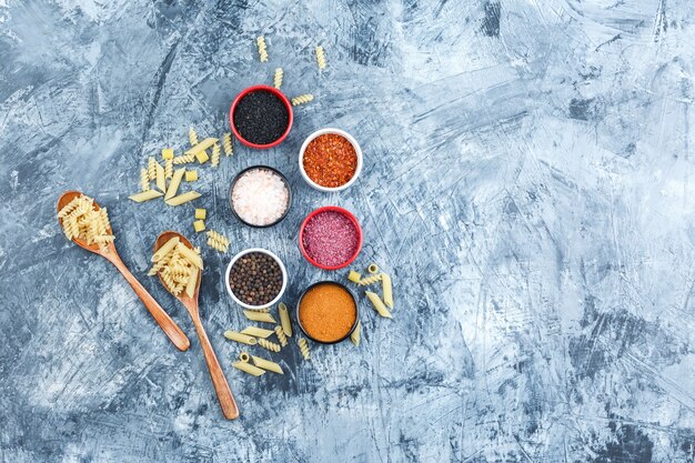 Raw pasta in wooden spoons with spices top view on a grey plaster background