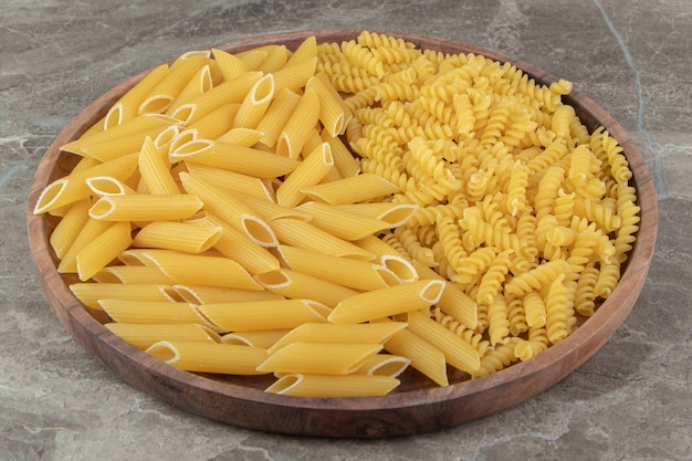 Raw penne and fusilli pasta on wooden plate.