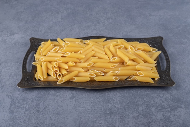 Raw penne pasta on dark tray.