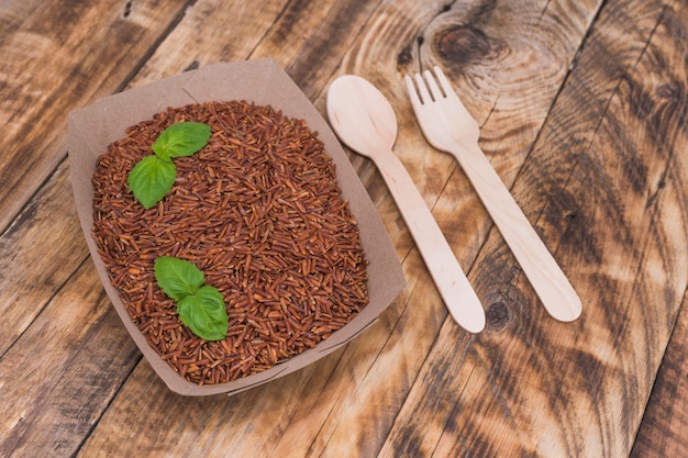 Raw red rice in container with basil leaves; spoon and fork over wooden surface