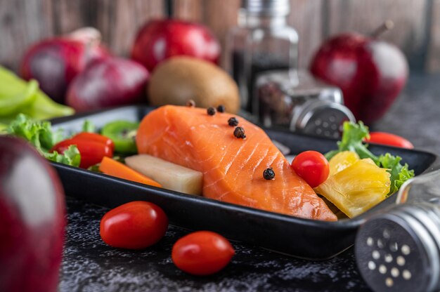 Raw salmon fillets, pepper, kiwi, pineapples, and rosemary onto a plate and black cement floor.