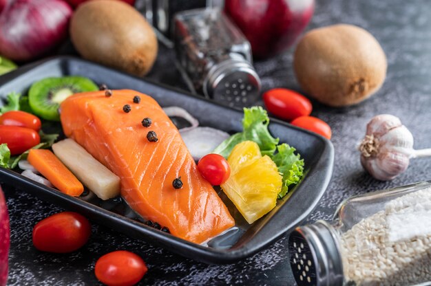 Raw salmon fillets, pepper, kiwi, pineapples, and rosemary onto a plate and black cement floor.