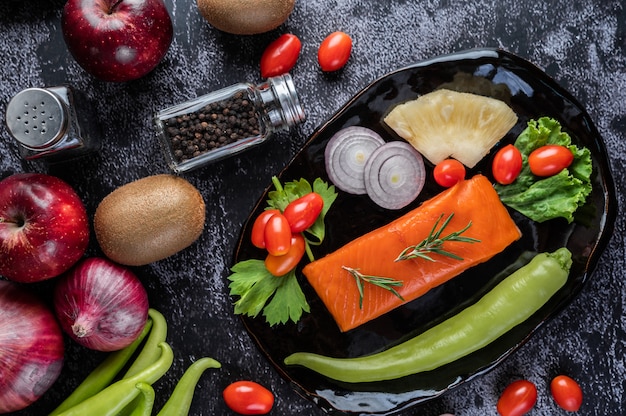 Сырые филе лосося, перец, киви, ананасы и розмарин на тарелку и черный цементный пол.