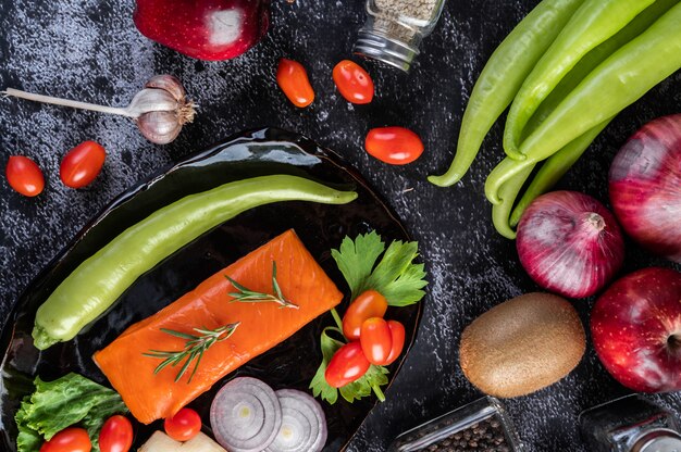 Raw salmon fillets, pepper, kiwi, pineapples, and rosemary onto a plate and black cement floor.