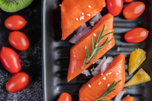 Raw salmon fillets, pepper, kiwi, pineapples, and rosemary onto a plate and black cement floor.