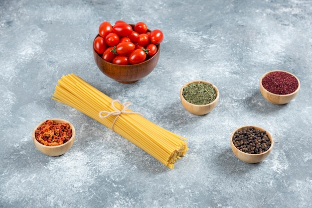 Free photo raw spaghetti, bowl of tomatoes and spices on marble background.