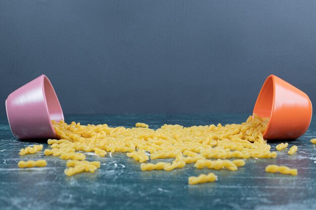 Raw spiral pasta in colorful bowls on blue space. 