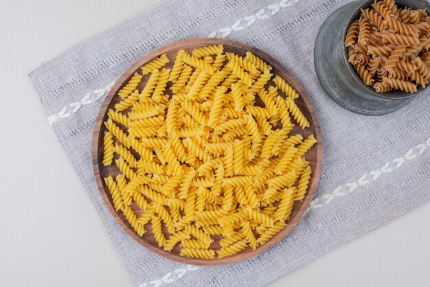 Raw spiral pasta in cup and plate on tablecloth.