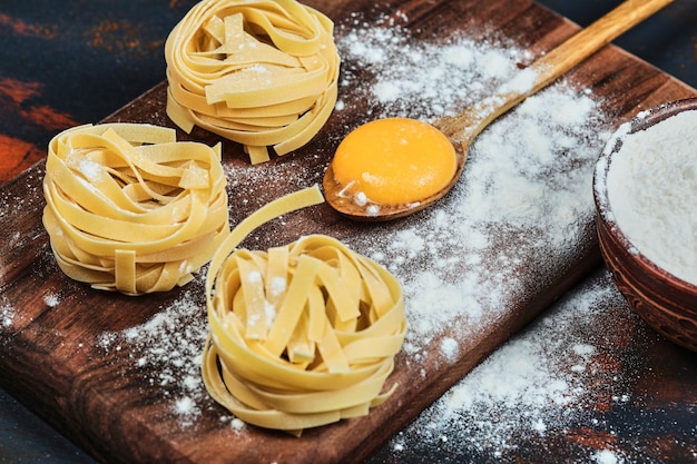 Raw tagliatelle pasta on wooden board with bowl of powder.