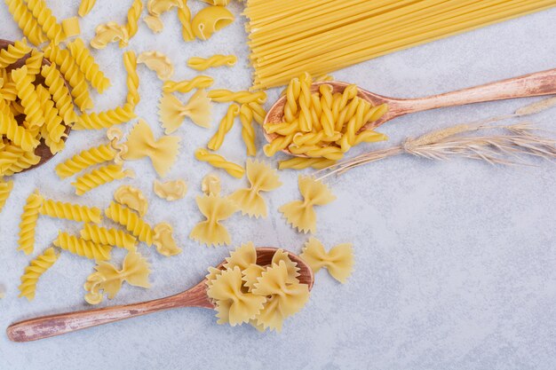 Raw various pasta shapes and spoons on white surface