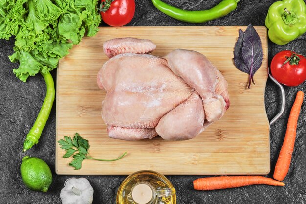 Сырая целая курица на деревянной доске с салатом, перцем, маслом и помидорами