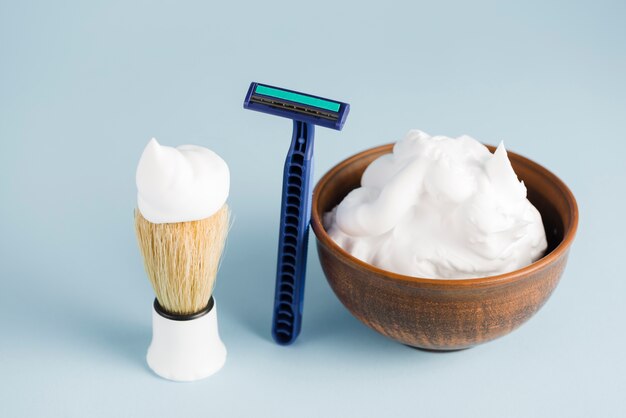 Razor; shaving brush and bowl of foam against blue backdrop