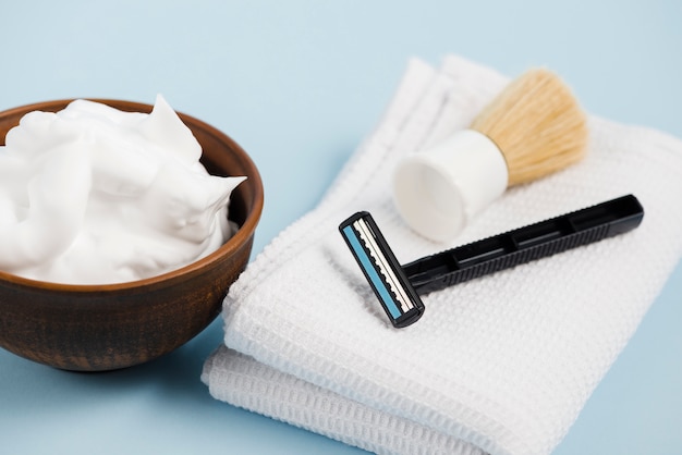 Razor and shaving brush over the folded napkin near the wooden bowl with foam against blue background