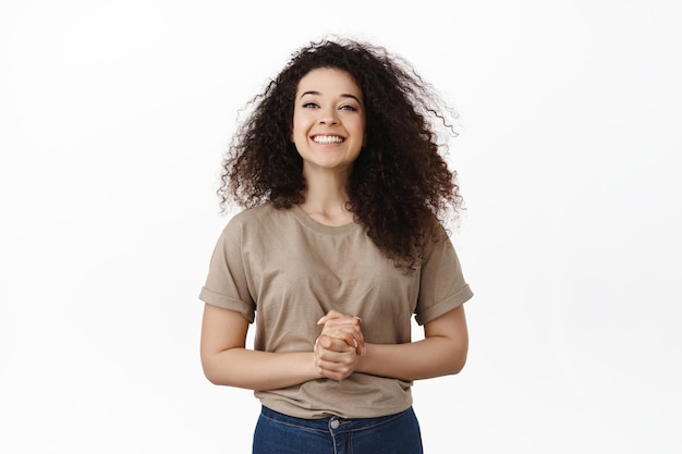 Free photo ready to assist you, how may i help. smiling friendly girl looking happy at camera, holding hands clenched together near chest, standing like assistant, answer your questions, white background