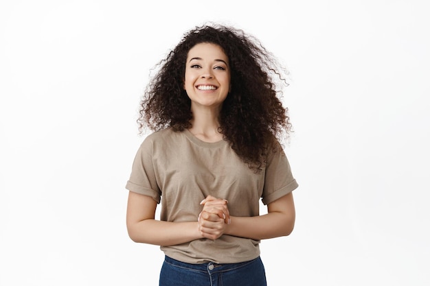 Ready to assist you, how may I help. Smiling friendly girl looking happy at camera, holding hands clenched together near chest, standing like assistant, answer your questions, white background