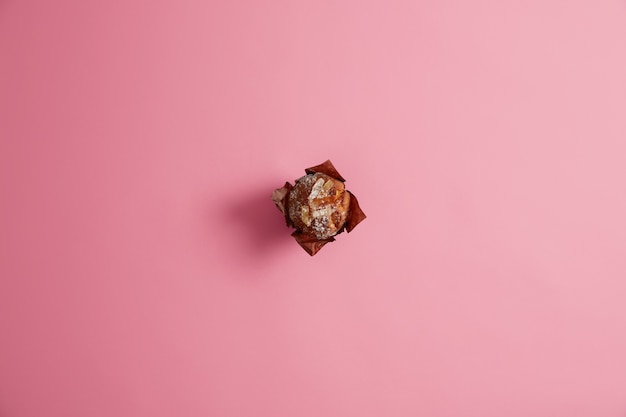 ピンクの背景に分離された茶色の紙に砂糖を粉末にした焼きたてのマフィン。新鮮な菓子、甘い生活、ジャンクフードのコンセプト。朝の朝食。グルメのデザート。セレクティブフォーカス。