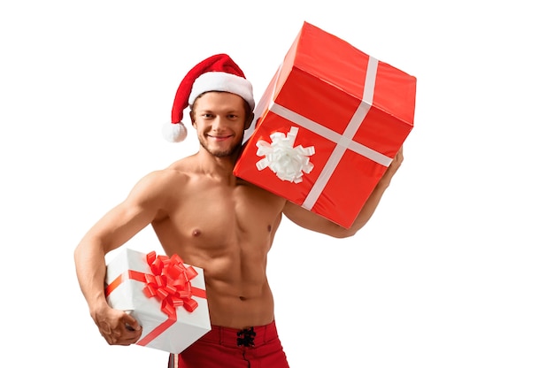 축하할 준비가 되었습니다. 그의 찢어진 몸을 보여주는 선물을 들고 웃는 산타 클로스 모자를 쓰고 섹시 한 매력적인 남자