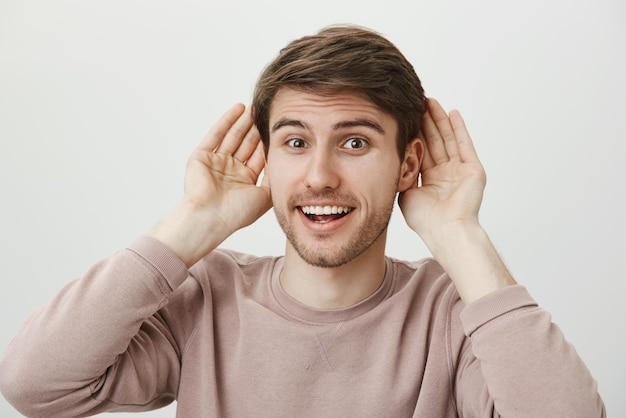 Free photo ready to receive new information studio portrait of attractive positive guy holding palms near ears and smiling happily hearing rumor or eavesdropping standing over gray background