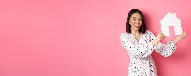 Free photo real estate beautiful asian woman demonstrating paper house model looking at camera confident advert