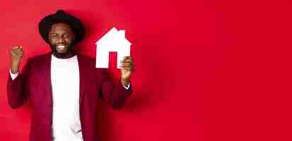 Free photo real estate cheerful black man rejoicing and showing paper home maket standing over red background