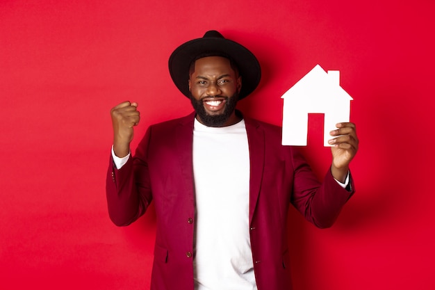 부동산. 쾌활 한 흑인 남자 기쁨과 빨간색 배경 위에 서있는 종이 홈 maket를 보여주는.