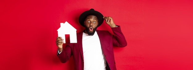 Real estate cheerful black man showing paper house and smiling recommending broker standing over red