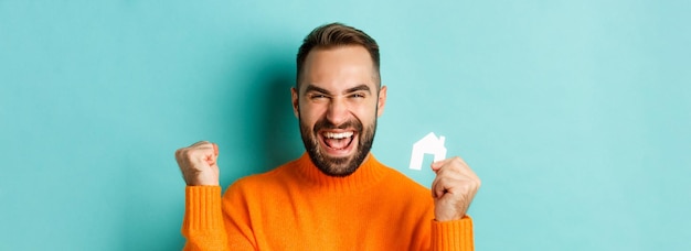 Real estate cheerful man buying apartment rejoicing and saying yes showing small paper house standin