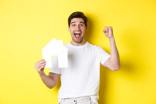 Концепция недвижимости. Веселый человек показывает модель бумажного дома и делает кулачковый насос, оплаченную ипотеку, желтый фон.