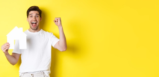 Free photo real estate concept cheerful man showing paper house model and making fist pump paid mortgage yellow