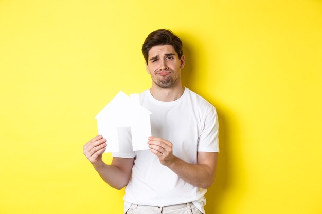 Real estate concept displeased young man showing paper house model and grimacing upset standing over...