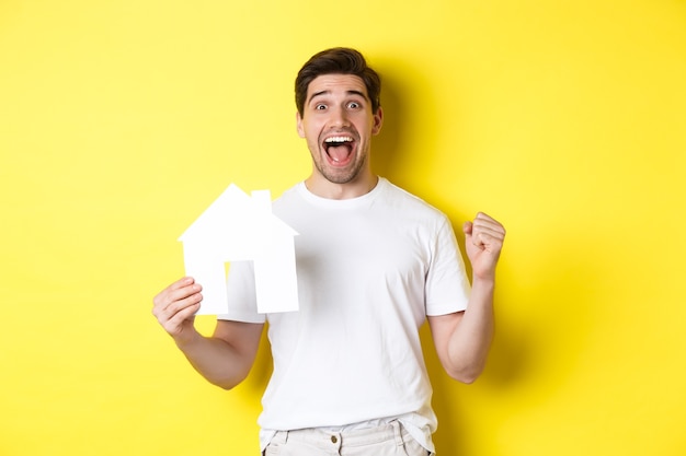 Real estate concept. Excited man holding paper house model and celebrating, standing happy over yellow background.