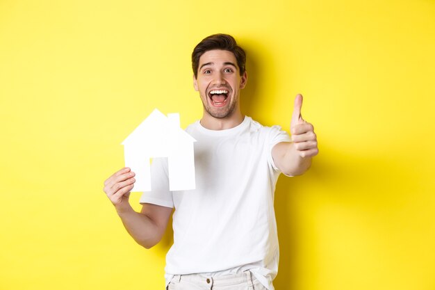 Концепция недвижимости. Счастливый молодой мужчина покупатель показывает палец вверх и бумажную модель дома, улыбаясь довольным, стоя на желтом фоне.
