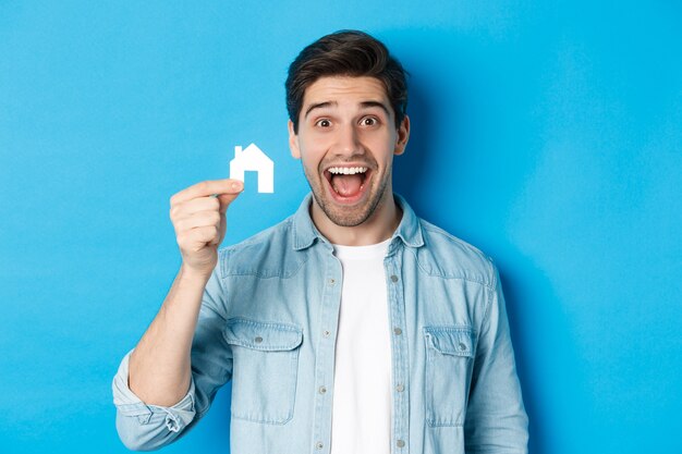 부동산 개념입니다. 행복해 보이는 젊은 남자는 파란색 배경 위에 서 있는 작은 집 모델을 보여주는 아파트를 찾았습니다.