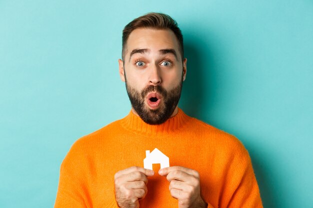 Real estate concept. Happy young man searching for home rent, holding  paper house and gasping