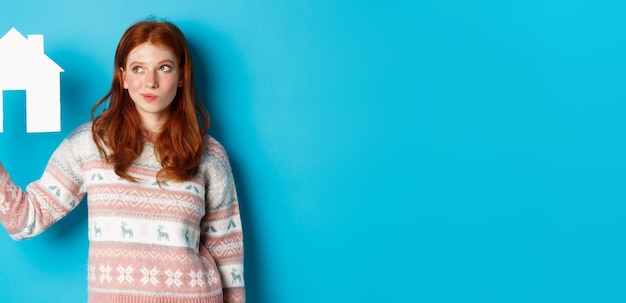 Real estate concept image of thoughtful redhead girl showing paper house model and thinking searchin