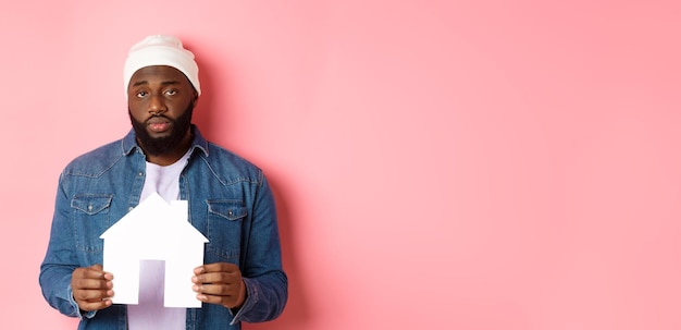 Foto gratuita concetto di bene immobile uomo di colore triste e stanco che fissa distratto alla macchina fotografica che tiene il modello di casa di carta sta