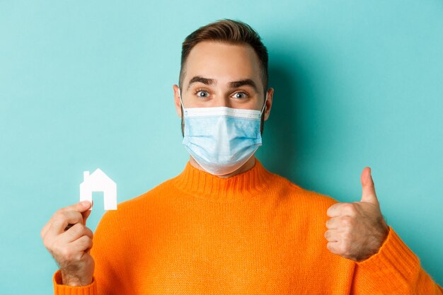 Real estate and coronavirus pandemic concept. Close-up of adult man in medical mask holding small house